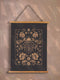 A symmetrical wall tapestry featuring a large scarab beetle in antique bronze tones surrounded by detailed flowers and botanical elements on a black background, mounted on wooden dowels with twine.